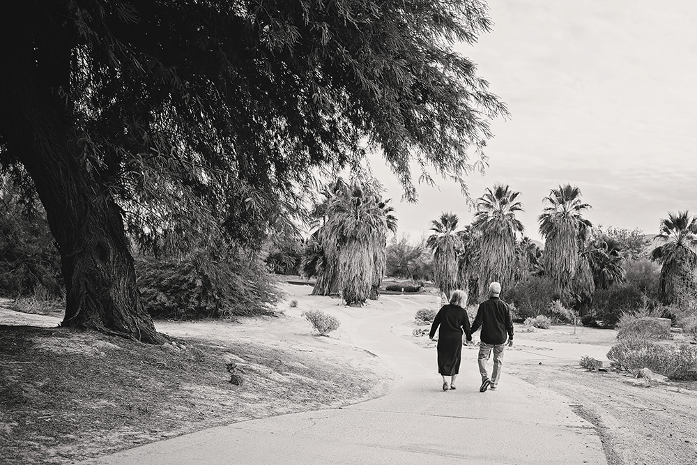 A black and white photo of an older couple strolling away from the camera amongst lush trees
