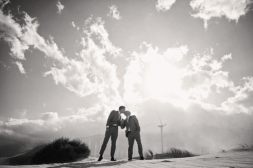 Black and white portrait of two grooms kissing outdoors with dramatic clouds, desert mountains, and wind turbines near Palm Springs after their Avalon Hotel wedding.