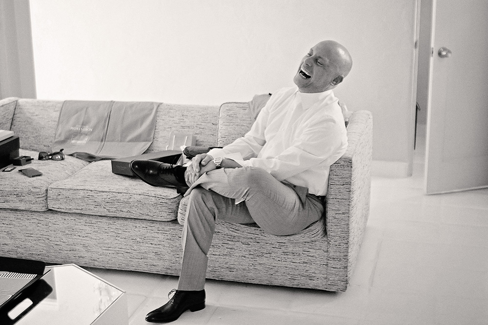 Black and white candid photo of a groom laughing while putting on his shoes during wedding preparations at the Avalon Hotel in Palm Springs.