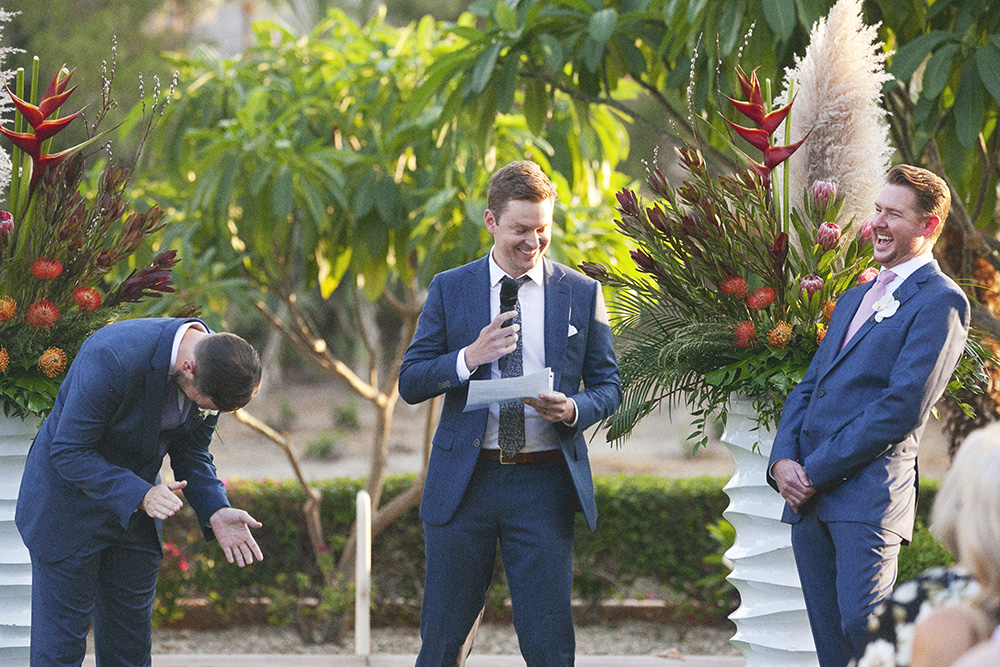 Groom laughing during heartfelt speech at same-sex wedding ceremony in Indian Wells California with tropical floral arrangements