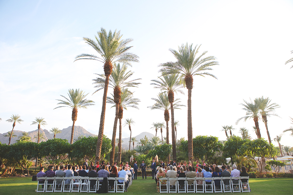 Outdoor same-sex wedding ceremony in Indian Wells, California with guests seated beneath tall palm trees and desert mountains in the background
