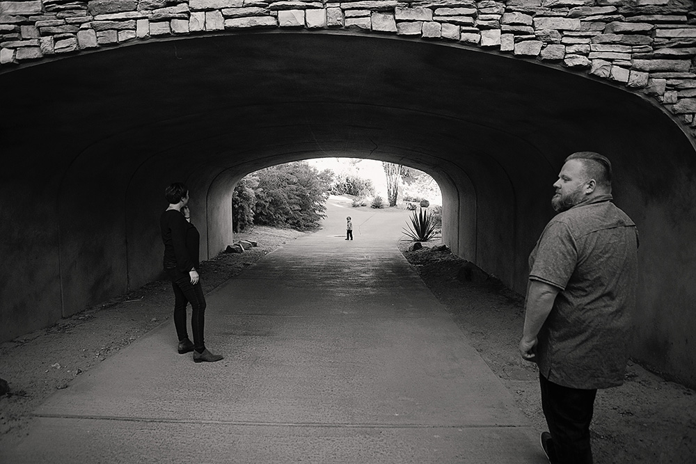 Toddler plays under a bridge as his parents look on